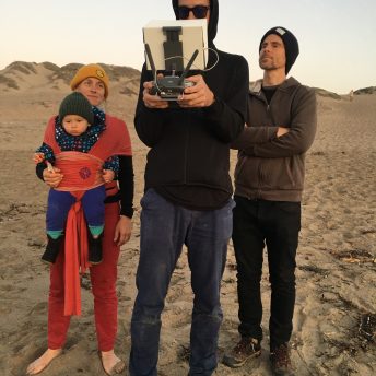 Three figures, one woman and two men, stand on a beach looking at the drone control held by the middle person. On the left is a woman with a baby dressed in red.