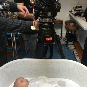 A small baby wrapped in white lies in an oval crib. A camera's lens is pointed directly down on him from above.