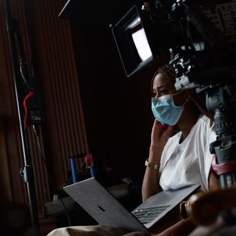 A Black woman with a medical mask on sits near a cinema camera.
