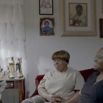 Two brown-skinned Black women smile while seated in a living room with various framed portraits and figurines.
