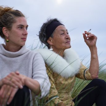 Director Lin Alluna and main character Aaju Peter sitting next to each other in a field, looking out in to the distance. Aaju is smoking a cigarette.