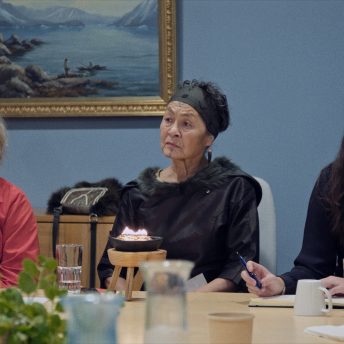The main character of Twice Colonized, Aaju Peter, is in a meeting and is sitting next to two young fellow Inuit activist. There is a traditional Greenlandic oil lamp, a qulleq, in front of her, and all three women are looking serious but hopeful.