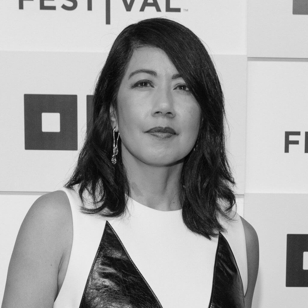 Black and white portrait of Beth Aala looking at the camera. She has light brown skin, and dark brown shoulder length hair.