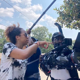 a collection of Black people in formal clothing stand around a projector screen smiling while a Black man in dark clothes points his camera toward them