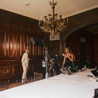 a Black woman in a dark jumpsuit laughs with an older Black man in a suit as he stands under a production light for an interview