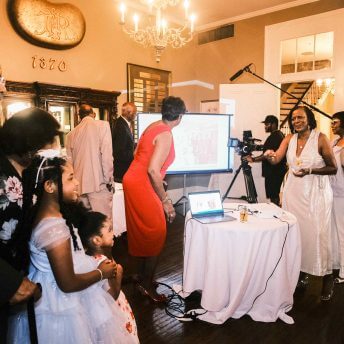 a collection of Black people in formal clothing stand around a projector screen smiling while a Black man in dark clothes points his camera toward them