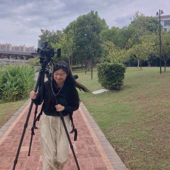 Viv, the director, carries an open tripod on her shoulders, mounted with camera and gears, while rushing down a riverside greenbelt.