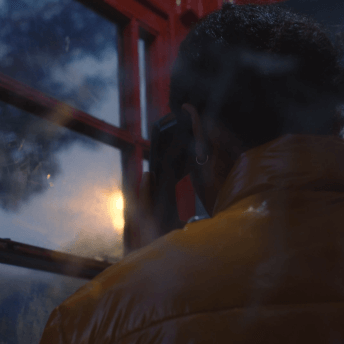 At dawn, a woman in a yellow puffer jacket holds a phone to hear ear in a red old-fashioned phone booth.